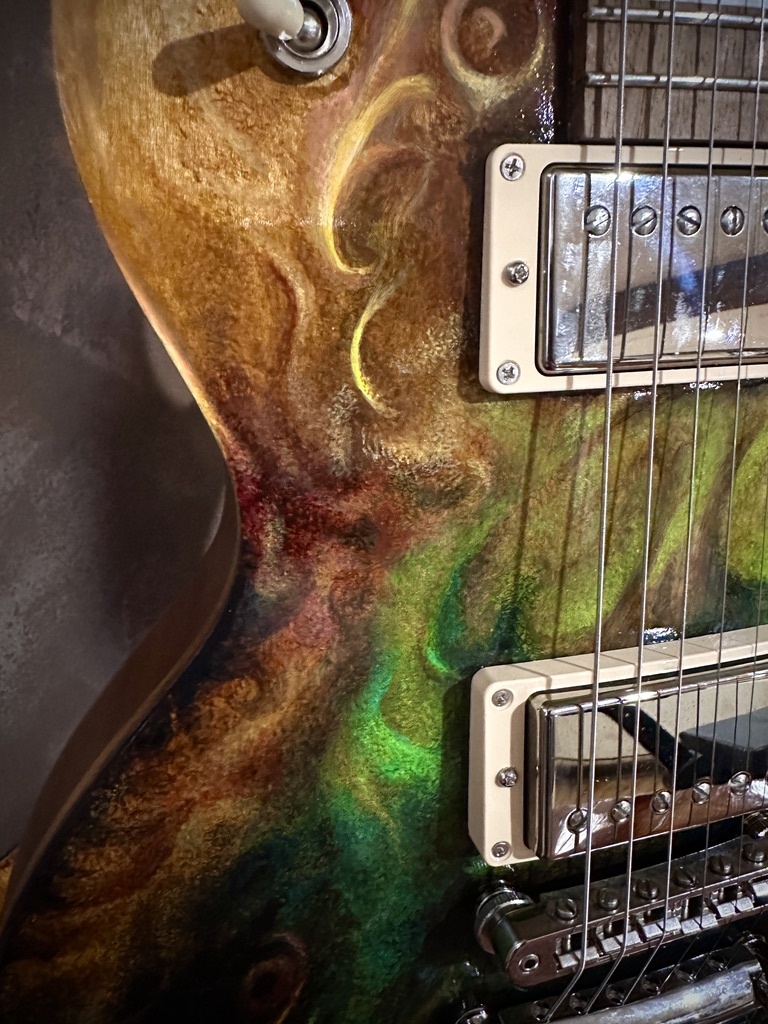 Close view of translucent gold, brown and green painting beside the pickups of an electric guitar.