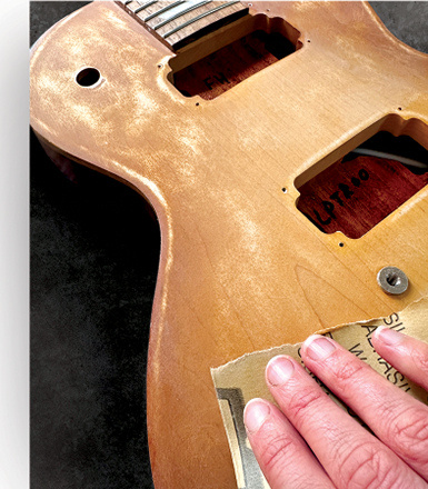 Hands preparing the surface of a guitar body at the worktable.